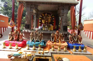 An outdoor shrine decorated for worship, with a tiered stone temple structure behind an altar. Multiple deity murtis, several dressed in white and gold, are arranged on coloured cloth bases in pink, teal, orange and blue, each garlanded with white flower strands. Brass kalashas with mango leaves and coconuts sit on stone plates in front, with offerings of laddus, rice, flowers and turmeric on a low table. Orange and red curtains frame the shrine pillars, and small decorative lanterns hang at the sides. No people are visible.