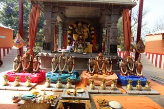 A temple shrine set up for a homa or puja, with a central deity murti of a seated couple draped in garlands at the back of a stone shrine, flanked by rows of smaller garlanded murtis of deities and sages standing on coloured cloths in red, teal, orange and blue. Brass kalashas with mango leaves and coconuts sit on a low platform in front, with bowls of sweets, rice, fruit and a fire pit at the lower edge. Orange curtains hang at the sides and orange and red decorative pennants hang from the pillars. No people are visible.