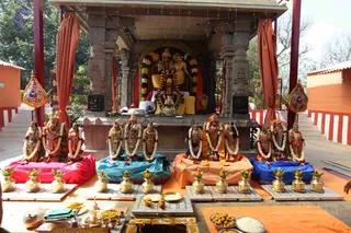 A photograph of a temple shrine set under a stone canopy with red-and-orange curtains hanging at the sides. At the centre, a seated deity murti pair is garlanded with white and yellow flower strings. In front, a row of about ten small deity murtis is arranged on coloured cloths of red, blue, orange and blue, each draped with white flower garlands. Brass kalasha pots with mango leaves and yellow flowers stand on the platform in front. Brass and silver ritual vessels, plates of sweets, rice and fruit occupy the foreground. No people are visible in the scene.