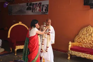 A woman in a red and green silk sari with a white blouse feeds a bald man in white ritual dress a sweet from her fingers. The man wears a white dhoti and shawl with gold borders and a long garland of white flowers and red roses, and has a red bindi on his forehead. Behind them stands an ornate gold-and-red throne-style chair and a gold carved sofa on an orange wall. A framed printed collage on the wall shows photographs of older men and a group of people, with a caption reading 'A Search with Essence' and names including Sri Ramana Maharshi and Sri Gurudev Bhagavan. Scattered flower petals and a mat lie on the floor.