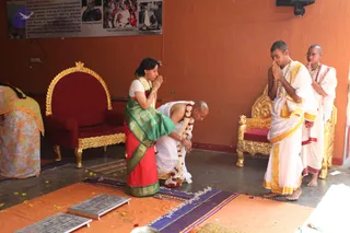 A photograph of a ritual greeting in an orange-walled hall. A woman in a red sari with a green and gold border and a white blouse, wearing a white marigold garland, kneels with palms pressed together while bowing before a man in white dhoti and shawl with a garland and a shaved head, who stands bent forward and holds her hands. Behind them, two young men in white dhotis with gold borders stand with palms joined in namaste; one wears a rudraksha mala and gold necklace. Two empty ornate gold-and-red thrones stand against the wall, and orange and purple mats with scattered yellow flower petals lie on the floor. A framed poster with photographs and text hangs on the upper wall.