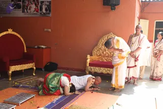 Indoor ritual scene in an orange-walled hall with a gold-carved red-cushioned throne and a red armchair at left. Two people prostrate on the floor in full padanamaskar on a saffron cloth and purple mat, scattered flower petals nearby. A woman in a red and green silk sari with a white flower garland lies prone, and a shaven-headed man in white dhoti with gold border lies beside her. A third man in white dhoti with gold border and a gold-beaded necklace bows low at the right. Two more men in white dhotis stand near a woman in a pink-and-white sari. A framed photo collage hangs on the wall above the chairs.