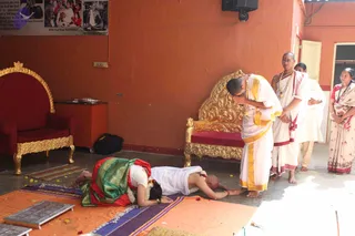 A photograph inside an orange-walled hall with gold-carved thrones and a red-cushioned seat. In the foreground, a woman in a green and red silk saree with a white blouse lies prostrate with her forehead to the floor on a purple-and-orange cloth, a flower garland around her neck and rose petals scattered nearby. Beside her, a bald man in white dhoti and shawl with gold trim bends forward to touch her head in blessing. Behind him, a man in white with a gold necklace and rudraksha-style beads stands with hands clasped, and a woman in a sari with a red border watches at the right. A small wall-mounted photo collage and a black bag sit in the background, and a flat ritual tray lies at the left edge.