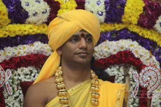 A single man seated before an elaborate backdrop of marigold, jasmine and rose garlands arranged in layered rows of yellow, purple, white and red. He wears a saffron-yellow turban, a yellow shawl draped over bare shoulders, a heavy gold necklace of large bead-like pendants and a fine gold chain, and a red kumkum mark on his forehead. A decorative gold and silver ornament is visible at the right edge. Appearance-based cues are consistent with a Swamiji (probable) seated in ceremonial honour, with turban, saffron clothing and ornate gold necklace.