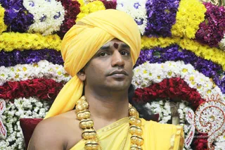 A close portrait photograph of a man seated in front of a floral backdrop of yellow, white, purple and red flowers arranged in tiers. He wears a yellow turban and yellow robe with a gold trim, a red tilak mark on his forehead, and a heavy ornate gold necklace of large beads. A decorative silver-and-red ornament appears at the lower right.