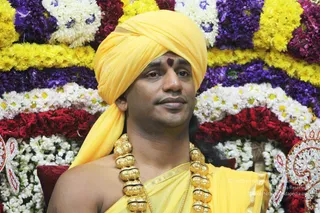 Close-up portrait of a bare-chested man seated before a backdrop of layered flower garlands in yellow, white, purple and red marigold-style blooms. He wears a bright yellow turban with a red tilak mark on the forehead, a yellow shawl draped over the shoulders and a thick ceremonial gold necklace of large beads. Ornate gold and red decorative elements frame the edges of the flower arrangement. The scene resembles a ceremonial or satsang setting.