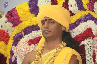 A photograph of a man seated in front of a large floral backdrop of marigolds and white, red and purple blooms arranged in a semicircular halo. He wears a yellow turban and yellow shawl, a red tilak on his forehead, long hair falling behind his shoulder, and heavy ceremonial gold beaded necklaces. A jewelled ornament with silver-toned stonework is visible at the lower left. Swamiji (probable): the candidate wears saffron-yellow ornate clothing, a turban, long swept-back hair and ceremonial gold necklaces, consistent with the page's reference to Swamiji at this event. He is the only physical person visible.