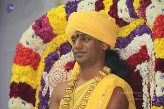 A man in a saffron-yellow turban and matching yellow silk drape sits before a backdrop of garlands in yellow, white, red and purple flowers. He wears a red tilak on his forehead and a heavy ornate gold necklace of large round beads over his bare shoulder. A sparkling jewelled ornament is visible at his left side. The composition is a close, waist-up portrait against the floral arch.