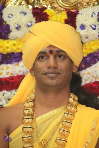 Close portrait of a man wearing a bright yellow turban and matching yellow shawl, with a red kumkum mark on his forehead. He wears heavy ornate gold beaded necklaces with dark accents over the shawl. Behind him is a wall of garlands in white, yellow, red and purple flowers with blue decorative accents. Swamiji (probable): long hair visible at the back, ceremonial gold necklaces and a yellow turban and shawl match the presentation described on the page.