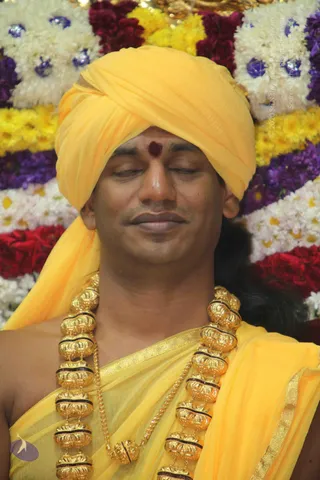 Close-up photograph of a man with eyes closed and a slight serene smile, wearing a bright yellow turban and matching yellow shawl over a yellow garment with gold border. A red tilak mark is on his forehead, and he wears heavy gold ceremonial necklaces with bead-like links. Behind him is a backdrop of white, yellow, red and purple flower garlands with blue accents. The appearance is consistent with a person turbaned figure in saffron-yellow ceremonial dress with gold ornaments, seated in a flower-decorated setting, apparently in meditation or darshan.