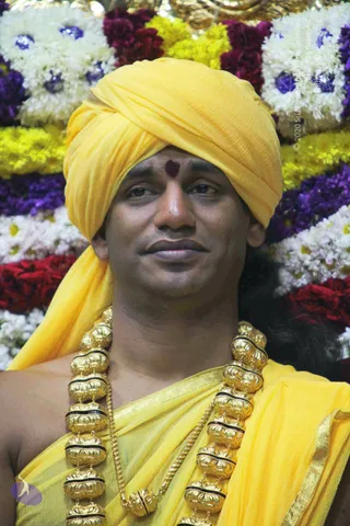 Close portrait of a man wearing a bright yellow turban and matching yellow shawl draped over bare shoulders, with a dark red tilak mark on his forehead. He wears a heavy ornate gold necklace of large beaded segments with black bands. Behind him is a dense backdrop of white, yellow, red and purple flower garlands with blue-centred blossoms.