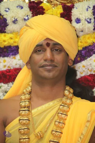 Close portrait of a man wearing a bright yellow turban and matching yellow shawl draped over bare shoulders. He has a red bindi on his forehead and wears a heavy ornate gold necklace of large beaded links with dark bands. Behind him is a backdrop of garlands of white, yellow, red and purple flowers with blue accents.