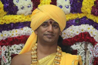 A close-up photograph of a man wearing a bright yellow turban and yellow-saffron garments, with a red kumkum mark on his forehead. Heavy gold ceremonial beads hang around his neck. Behind him is a backdrop of garlands of white, yellow, red and purple flowers, with decorative silver-and-red ornaments at the edges. Only one person is visible.