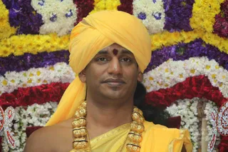A close portrait photograph of a man wearing a bright yellow turban and matching yellow cloth draped over his bare shoulder, with a red tilak mark on his forehead. He wears heavy gold ceremonial beaded necklaces. Behind him is a dense floral backdrop of yellow, white, purple and red flowers arranged in horizontal bands, with a sparkling decorative ornament at the lower edges.