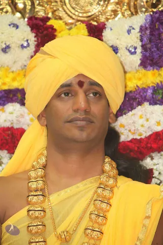 Close-up photograph of a man wearing a bright yellow turban and yellow silk shawl, with a red tilak mark on his forehead and a heavy gold ornamental necklace of large fluted beads. Behind him is a backdrop of white, yellow, purple and red flower garlands with a gold ceremonial element at the top. A single person is shown, looking to the side with a composed expression. Swamiji (probable): the saffron-yellow turban and shawl, gold necklace and long hair are visible cues, but the identification is based on appearance only.