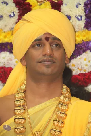 Close-up photograph of a man wearing a bright yellow turban and a matching yellow shawl draped over his shoulders. A reddish-maroon tilak mark is on his forehead, and he wears heavy ornate gold necklaces with dark bead separators. Behind him is a backdrop of white, yellow, red and purple flower garlands. He gazes calmly to the side.