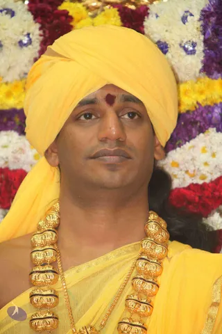 Close-up photo of a man wearing a bright yellow turban and yellow robe with a red tilak mark on his forehead. He wears a heavy multi-strand gold necklace of large ornate beads over the yellow cloth. Behind him is a backdrop of white, yellow, red and purple flower garlands. The man looks forward with a calm expression.