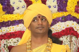 A close-up photograph of a man wearing a bright yellow turban and matching yellow silk garment, with a red kumkum mark on his forehead and heavy gold ceremonial necklaces around his neck. He looks off to the side. Behind him is a backdrop of densely arranged flowers in yellow, purple, white and red, with a glimpse of silver ornamental trim at the lower edges. Swamiji (probable): the long swept-back hair, yellow turban, saffron-yellow clothing and ornate gold necklaces match the appearance cues for the page's subject.