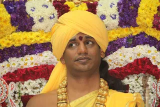 A man seated in front of a dense backdrop of flower garlands in yellow, white, purple and red marigold and jasmine-style blooms. He wears a bright yellow turban and yellow silk shawl, a red-brown tilak mark on his forehead, and a heavy gold ceremonial necklace of multiple gold beads. He looks off to the side with a composed expression. The Swamiji (probable) cues here are the yellow turban, long hair at the back, ornate gold necklace and saffron-yellow ceremonial clothing, shown as a single seated person at a satsang-style event.