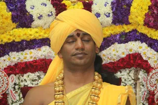 A close photo of a man with eyes closed in meditative stillness, wearing a bright yellow turban and yellow-orange shawl draped over his shoulders. A red tilak mark sits on his forehead, and he wears heavy gold ceremonial necklaces of large beads. Behind him is a dense floral backdrop of yellow marigolds, purple and white flowers, red roses and white blossoms arranged in bands, with ornate red and silver decorations at the edges.