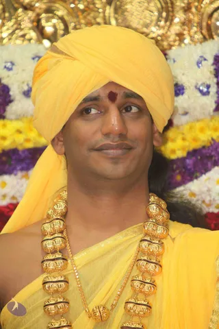 Close-up photograph of a man wearing a bright yellow turban and yellow draped garment, with a red tilak mark on his forehead. He wears heavy ornate gold beaded necklaces with a thin gold chain. Behind him is a backdrop of white, yellow, purple and red flower garlands. He glances to the side with a slight smile.