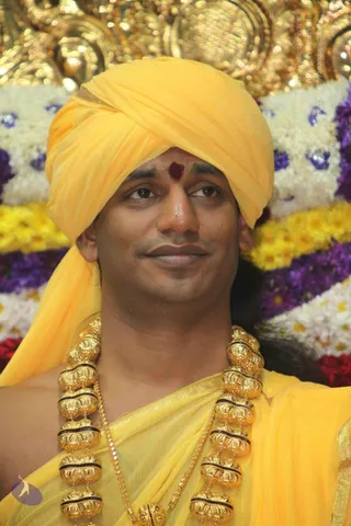 A close-up photograph of a man wearing a bright yellow turban and yellow silk garment, with a red tilak mark on his forehead and heavy ornate gold necklaces with dark beaded sections. Behind him is a backdrop of white, yellow and purple flower garlands and gold decorative elements. He has a slight smile and gazes off to the side. Swamiji (probable): the figure wears distinctive ceremonial gold necklaces and a saffron-yellow turban and garment within a flower-decorated setting.