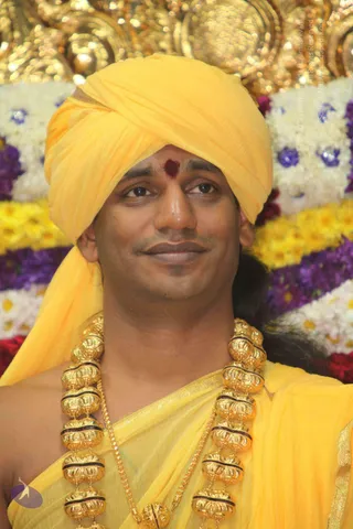 Close portrait photo of a man in a bright yellow turban and yellow silk wrap, with a red tilak on his forehead and heavy gold ceremonial beads around his neck. Behind him are garlands of white, yellow and purple flowers and a gold-toned decorative backdrop. Single person visible, smiling calmly.