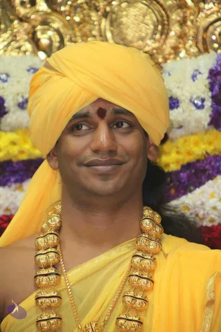 Close portrait of a man wearing a bright yellow turban and a matching yellow shawl over his shoulders, with a red bindi-style tilak on his forehead. He wears a heavy ornate gold necklace of large hollow beads with dark bands, and a long dark braid falls behind his shoulder. He gazes sideways with a slight smile. Behind him is a backdrop of gold ornamental decoration above rows of white, yellow, purple and red flowers.