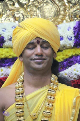 Close-up photograph of a man with eyes closed and a faint smile, wearing a bright yellow turban and yellow draped garment with gold border. He has a dark red tilak on his forehead and wears a heavy gold ceremonial necklace of large carved beads with black bands. Behind him are rows of white, yellow, purple and red flower garlands and an ornate gold backdrop.