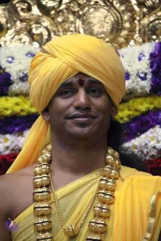Close portrait photo of a smiling man wearing a bright yellow turban and yellow draped garment, with a red bindi on his forehead and a heavy gold necklace of large ornate beads with black bands across his chest. Behind him are rows of white, yellow and purple flowers and a gilded ornamental backdrop.