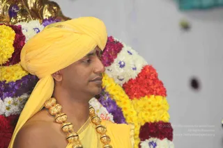 A man in profile wears a yellow turban and a yellow shawl draped over bare shoulders, with a red tilak mark on his forehead and a heavy gold bead necklace. He stands in front of a semicircular backdrop of marigolds and chrysanthemums in yellow, red, white and purple, with a gold ornamental frame at upper left. The scene appears to be a satsang setting.