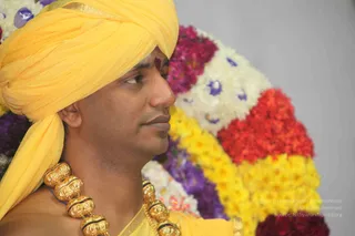 A close profile photograph of a man wearing a bright yellow turban and yellow garment, with a red tilak mark on his forehead and a heavy ornate gold necklace of large carved beads with black bands across his chest. Behind him is a large floral arrangement of white, yellow, red and purple flowers, with purple flowers pinned in the white blossoms. The background is plain grey.