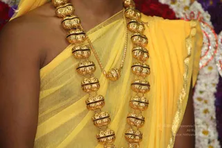Close-up photograph of a person's bare-shouldered torso draped in a sheer yellow cloth with a gold-thread border. Two long gold-toned bead necklaces with dark red-brown bands hang across the chest, joined by a thinner gold chain. Blurred red, white and purple floral garlands form the background. The face is not visible, so no individual can be identified and a person is unsure. Only the upper body of one person is visible.