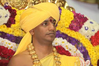 A man seated before a backdrop of densely arranged garlands of yellow, white, red, magenta and purple flowers. He wears a bright yellow turban and yellow garment draped over his shoulder, a red vermilion mark on his forehead, and a heavy ceremonial gold necklace of large beads with black accents. His gaze is directed off to the side with a calm, serious expression. The image is a close-up portrait of a single seated person in a flower-decorated setting.