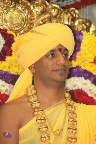 A man in a yellow turban and yellow silk garment stands in front of a backdrop of flower garlands in yellow, purple, red and white. He wears a large ornate gold necklace of beaded gold pieces with a fine gold chain, and a red tilak mark on his forehead. He looks off to the side with a calm expression. The gold backdrop behind him is ornamental.