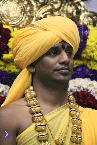 Close-up photograph of a man in a bright yellow turban and yellow shawl, with a dark red tilak mark on his forehead. He wears a heavy gold necklace of large ornate beads with black bands. Behind him are dense arrangements of yellow, purple, white and red flowers and a gold decorative backdrop.