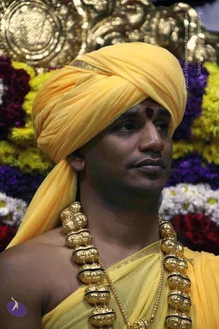 Close-up photo of a man in a bright yellow turban and matching yellow shawl draped over bare shoulders. He wears a heavy gold necklace of large ornamental beads with black accents and a fine gold chain, and a dark red mark on his forehead. Behind him is a dense arrangement of yellow, purple, white and red flowers with a gold ornamental backdrop. Appearance-based cues of a yellow turban, ornate gold beads and yellow shawl suggest a person in person, though the identification is uncertain.