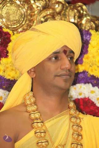 Close-up photograph of a man in a yellow turban and yellow garment, with a red kumkum mark on his forehead and a heavy gold necklace of large ornamental beads. Behind him are rows of yellow, purple, white and red flowers and a gold ornate backdrop. a person: the bare-shouldered figure wears a yellow turban and ornate gold necklaces, cues the page places on the a person album, but the face is not identifiable with confidence here.