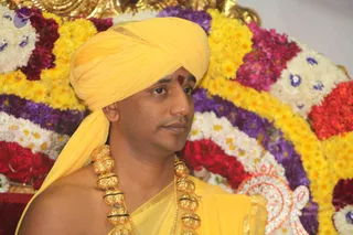 A close photograph of a man seated in front of a large flower backdrop arranged in bands of yellow, white, magenta, purple and red marigold and chrysanthemum garlands with blue accents. He wears a yellow turban, a red tilak on his forehead, a yellow shawl draped over bare shoulders, and a heavy gold necklace of large ornate beads. The appearance is consistent with a Swamiji (probable) candidate, supported by the turban, saffron-yellow ceremonial clothing and gold beaded necklace; he is shown in person. A partial circular sequined emblem is visible at lower right.