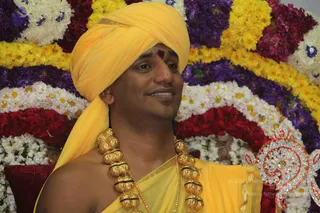 Close portrait of a man in a yellow-orange turban and matching yellow-orange robe, wearing a large gold ceremonial necklace of bead-like links and a red tilak on his forehead. He sits smiling slightly in front of a rounded arch of flower garlands in yellow, white, purple and red, with a red cushion and an ornate silver-and-red decorative piece at lower right. Swamiji (probable): the yellow-orange turban and robe, ornate gold necklace and seated ceremonial setting match the page's reference, though this is an appearance-based label only.