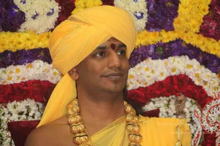 A man wearing a saffron-yellow turban and matching yellow draped garment sits in front of a dense backdrop of flower garlands in white, yellow, purple and red blooms. He has a red kumkum mark on his forehead and wears a heavy gold ceremonial necklace of large beaded links with black accents. His bare shoulder is visible and he gazes off to the side with a faint smile. A crystal-embellished decorative element appears at the right edge.