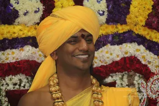 A smiling man in a yellow-orange turban and yellow shawl sits before a backdrop of densely arranged flower garlands in white, yellow, purple and red marigold and chrysanthemum strands. He wears a heavy gold ceremonial necklace of large knobbed beads and a red kumkum mark on his forehead. The photo is a close, cropped portrait of a single person seated in front of the floral decoration.