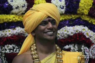 A smiling man in a saffron-yellow turban and matching yellow shawl sits before a backdrop of layered flower garlands in white, yellow, purple and red. He wears a large ornate gold necklace with a red-and-gold bead section and a red tilak mark on his forehead. The setting suggests a devotional gathering, and he appears to be receiving honour. Swamiji (probable): the saffron turban, shawl and ornate gold necklace are consistent with the page's description of the event, so this is an in-person candidate with a recognizable appearance.
