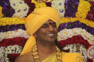 A smiling man in a saffron-yellow turban and yellow cloth draped over his bare shoulder sits before a backdrop of densely arranged flowers in yellow, white, purple and red. A red-orange tilak mark is on his forehead, and a heavy gold ceremonial necklace hangs on his chest. The bare-shouldered yellow wrap and turban resemble attire associated with a person, but the face and build are not used to confirm identity, so the appearance is only partially consistent.