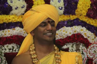 Close-up photo of a smiling man seated before a backdrop of densely arranged marigold, white jasmine and red and purple flower garlands. He wears a bright yellow-orange turban, a red tilak mark on his forehead, a saffron-yellow shawl draped over bare shoulders, and a heavy gold ceremonial necklace of large bead-like links. Glimpses of an ornate silver-and-red jewelled decoration appear at the lower right. Probable Swamiji by appearance, as the page identifies this event as a satsang featuring him; the turban, saffron clothing, gold necklace and tilak are the qualifying cues, shown seated in a garlanded setting.
