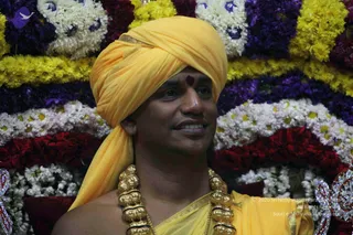 A close portrait of a smiling man wearing a bright yellow-orange turban and a matching yellow-orange shawl draped over his shoulders. He has a red tilak mark on his forehead and wears a heavy ornate gold necklace of large beads. Behind him is a backdrop of garlanded flowers in yellow, white, purple and red arranged in rows. Appearance-based label: Swamiji (probable), wearing a saffron-yellow turban, ornate gold necklace and yellow-orange shawl, seated in front of a flower decoration.