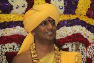 A man in a bright yellow turban and yellow shawl sits before a backdrop of flower garlands in yellow, white, purple and red. He wears a heavy gold ceremonial necklace of large beads and a red tilak mark on his forehead, and he is smiling and looking to the side. The appearance shows a long-haired, turbaned figure in saffron-yellow ornate clothing with gold necklaces, which fits the probable Swamiji cues, so the candidate is labelled Swamiji (probable) and present in person; the flower-decorated seat suggests a satsang or ceremonial setting.