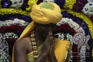 A close photograph of a man in a yellow turban and yellow draped garment, with his palms pressed together in anjali mudra and his eyes closed in a gesture of greeting or devotion. He wears a heavy gold necklace of large bead-like segments on his chest. Behind him is a circular arrangement of garlands in yellow, purple, white and red flowers, with a jeweled silver ornament on the right side.