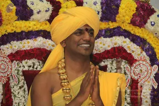 A smiling man in a yellow turban and yellow sash sits before a floral backdrop of yellow, white, purple and red flowers arranged in arches. He wears a red tilak on his forehead and a heavy ornate gold necklace of beads, and he holds his hands together in namaste. Silver and red jeweled decorations flank the floral arch. Swamiji (probable): the figure wears a yellow turban, a yellow sash and an ornate gold necklace, seated in a place of honour before a flower-decorated arch.
