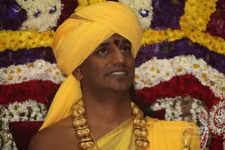 A man wearing a bright yellow turban and yellow silk garment with a gold-toned ornate necklace of large beads sits before a backdrop of densely arranged flowers in white, yellow, purple and red. A red tilak mark is on his forehead. He is turned slightly to the side and looks upward while speaking. a person: his long swept-back hair is not visible, but the yellow turban, yellow robes and ceremonial gold necklace match the page's claim of his presence, though the appearance cues are limited, so this is uncertain. Flowers and a jeweled ornament appear at the right edge.
